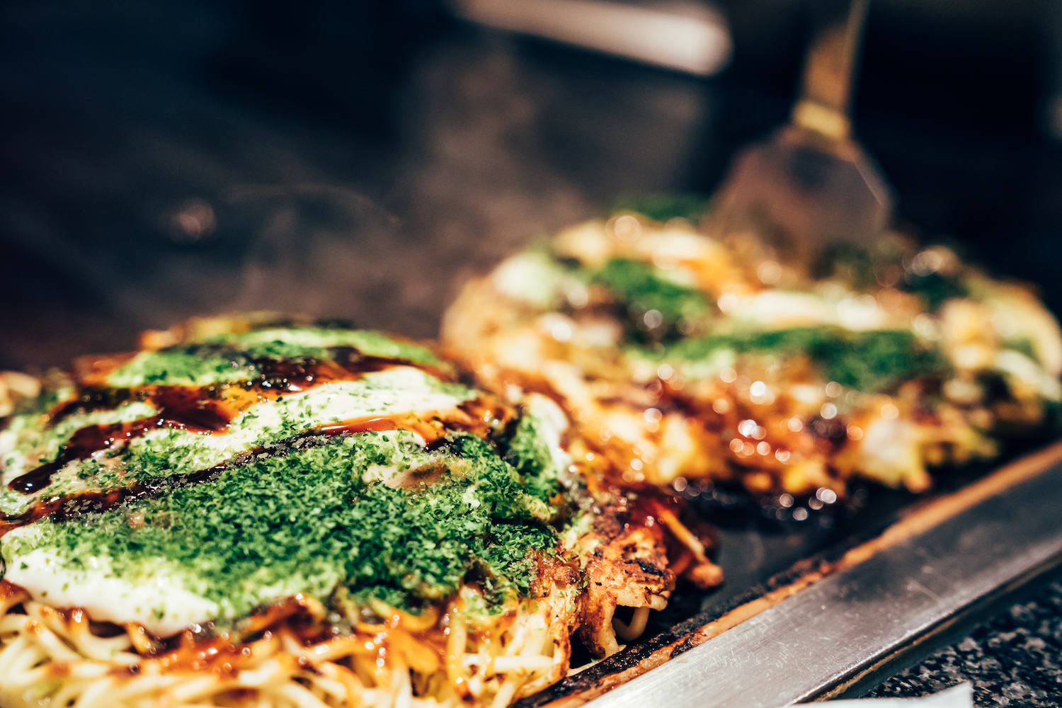 cooking okonomiyaki in local restaurant on teppanyaki pan. delicious Japanese hot plate pizza with green vegetable and sauce on it surrounding by smoke on iron plate indoor.